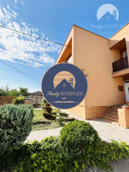 A family house in Okoč with a front garden and landscaped greenery in sunny weather.