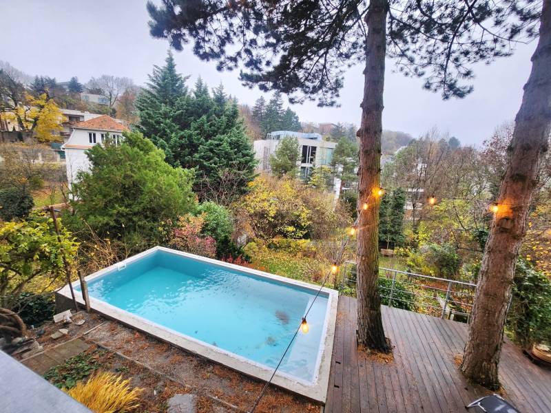 A wooden terrace with a pool surrounded by greenery and a view of houses in Bratislava.