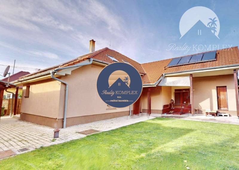 A family house in Komárno surrounded by a green lawn and paving under a blue sky.