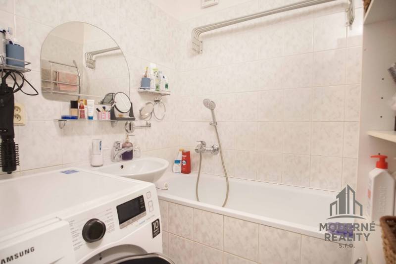A bathroom in a 3-room apartment with a washing machine, bathtub, and toiletries.