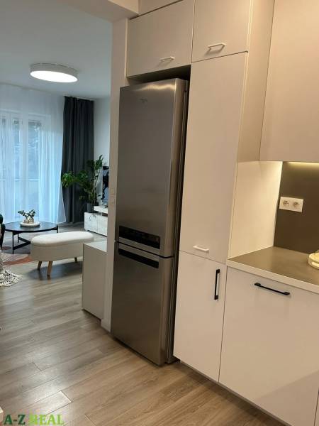 The kitchen of a 3-room apartment with a white kitchen unit, refrigerator, and wooden decor flooring.