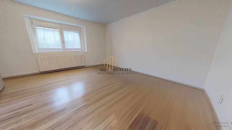 A room with white walls, a large window, and a wooden-patterned floor in a family house.