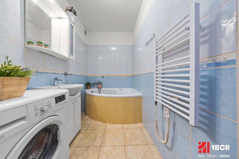 A bathroom in a 2-room apartment with a corner bathtub, a washing machine, and light blue tiles.