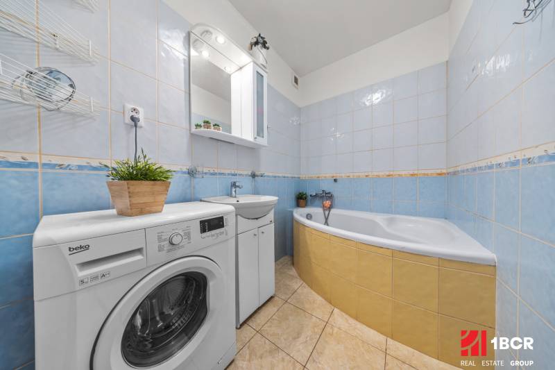 A bathroom with a bathtub, a washing machine, and light tiles in a 2-room apartment.