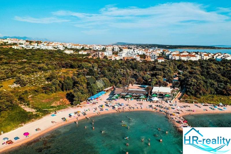 Beach with people, greenery, and 3-room apartments in the city of Novalja.