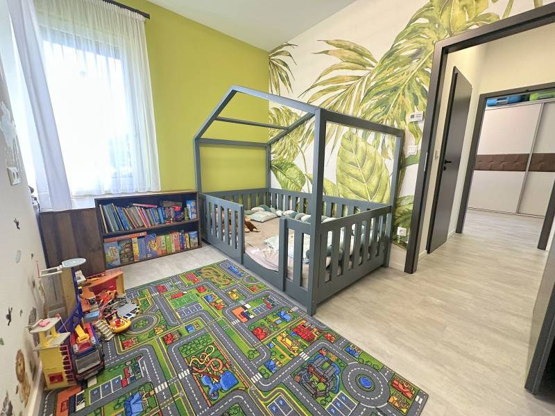 Children's room with a house-shaped bed, patterned carpet, bookshelf, and plant wallpaper in a family house.