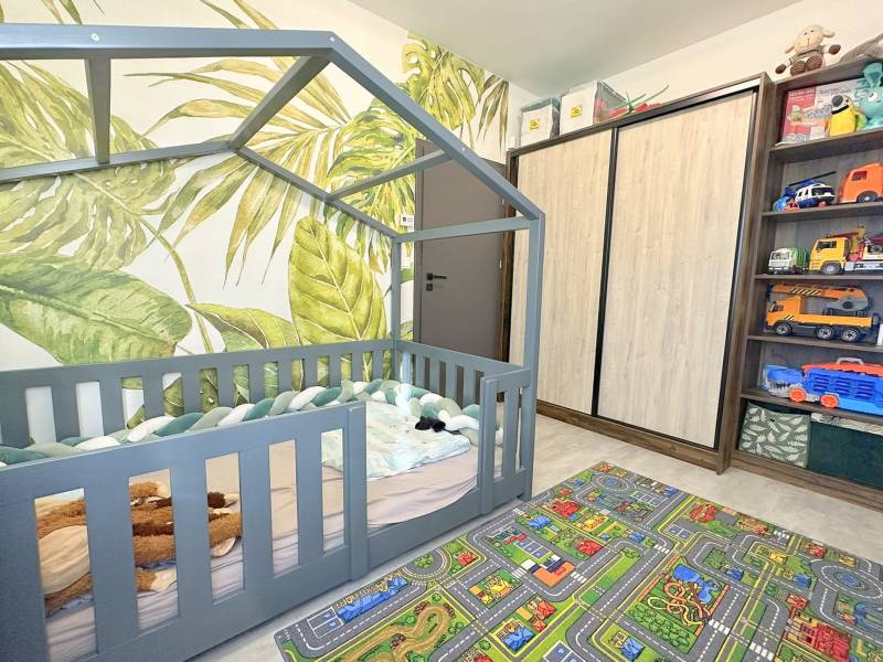 Children's room with a house-shaped bed, leaf wallpaper, and a play carpet.