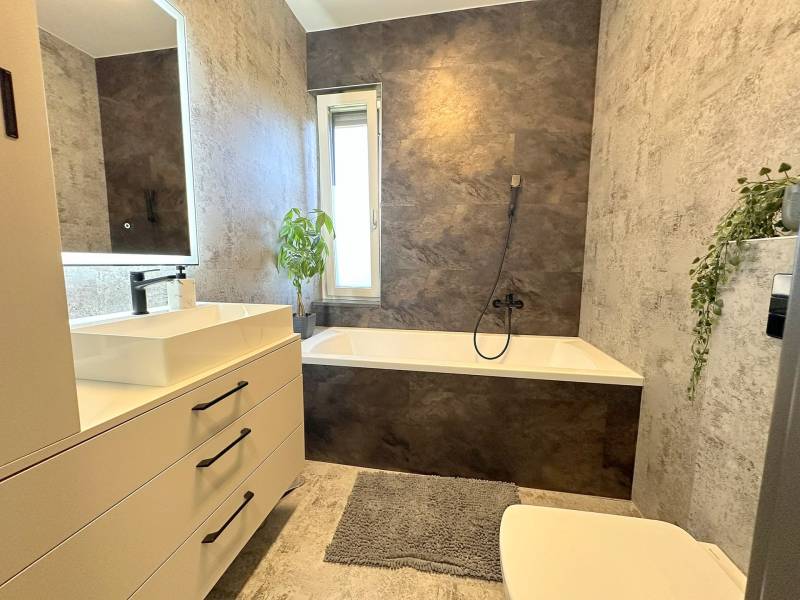 A bathroom in a family house with a bathtub, sink, plants, and tiled walls.