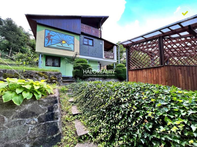 A cottage in Dudince with a colorful facade, a covered terrace, and a landscaped garden full of greenery.