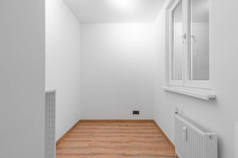 A white room with a window, a wooden decor floor, and a radiator in a studio apartment.
