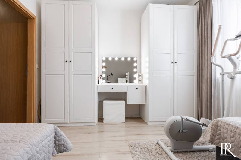A bright room in a 2-room apartment with a vanity table, white wardrobes, and an exercise machine.