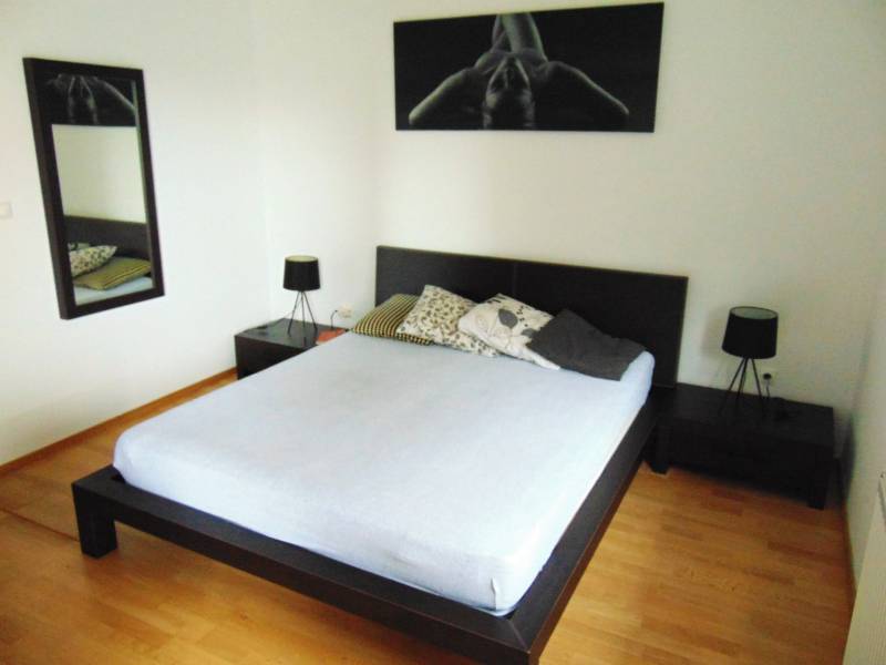 Bedroom of a one-room apartment with a double bed, mirror, and wooden decor flooring.