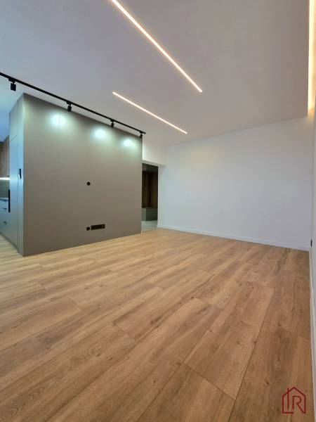A room with a wooden decor floor, lighting, and a kitchen area in a 3-room apartment.