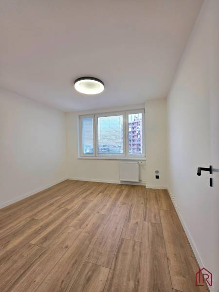 A room in a 3-room apartment with a wood-patterned floor and a window with a radiator.