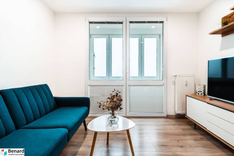 Living room in a two-room apartment with a sofa, television, and wooden decor flooring.