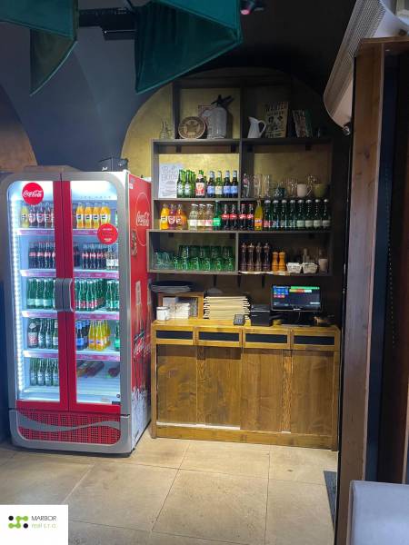 Gastro premises with a Coca-Cola refrigerator, shelves with drinks, and a cash register on a floor with a wooden decor.
