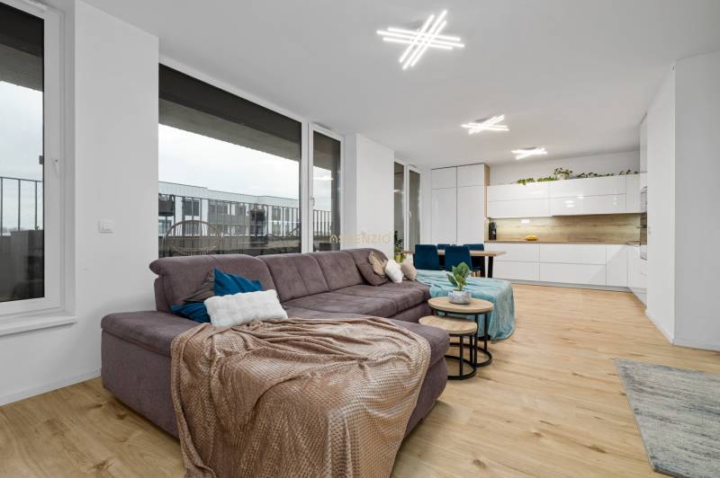 Living room with a brown sofa, white kitchen, and wooden decor flooring in a 3-room apartment.