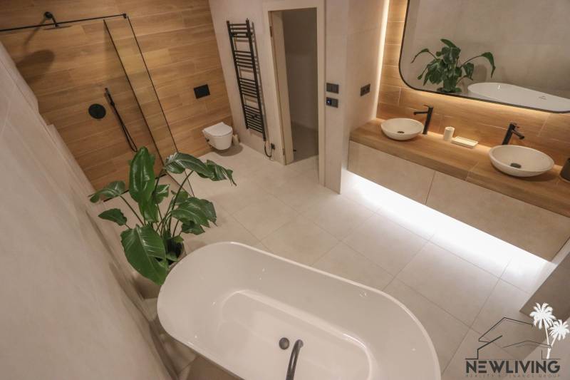 A bathroom in a family house with a bathtub, double sink, wood-patterned floor, and a plant.