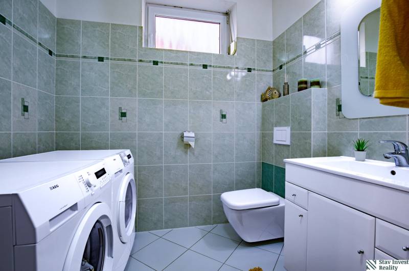 A bathroom in a family house with a washing machine, dryer, sink, and toilet, green tiles.