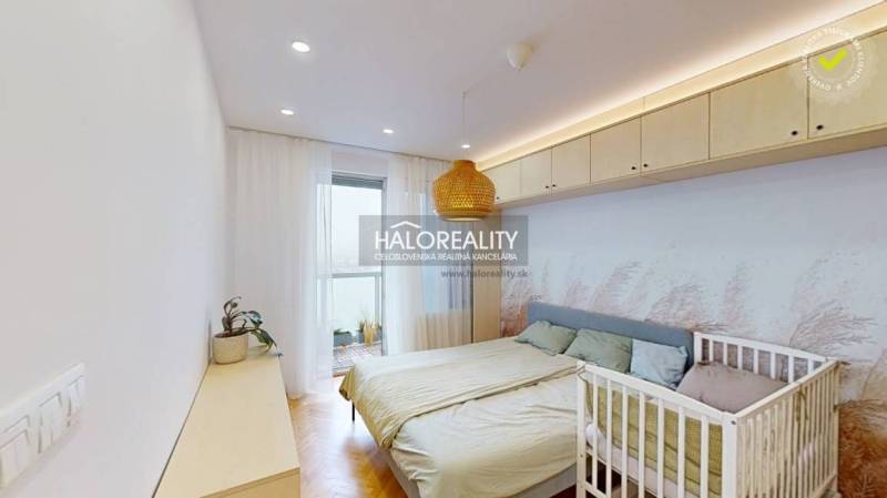 A bright bedroom with wood-patterned flooring, a double bed, and a baby crib in a three-room apartment.