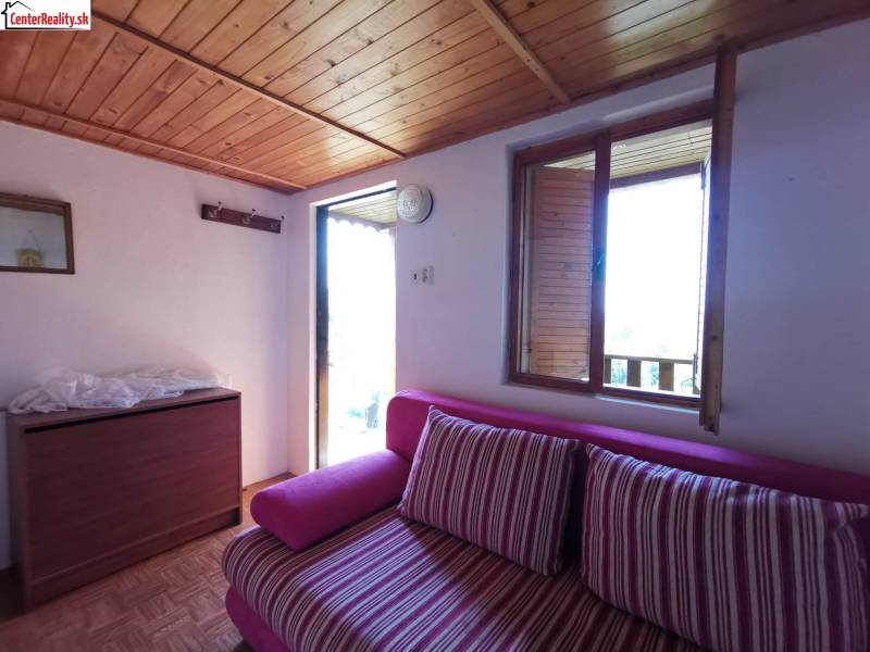 A room in a cabin with a pink sofa, wooden ceiling, and a view from an open window.