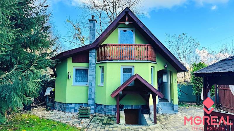 A cottage in Partizánske with a wooden balcony, surrounded by greenery and a paved courtyard.