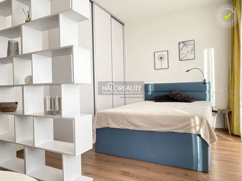 Interior of a 1-room apartment with a bed, white shelving unit, and wood-patterned flooring.