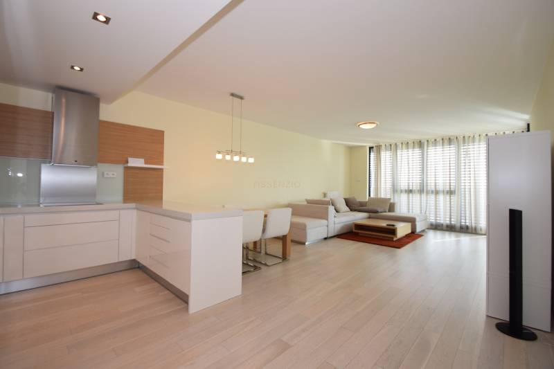 The living room of a three-room apartment with a wooden decor floor, a kitchenette, and large windows.