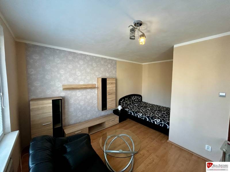 A room in a one-room apartment with a bed, a black armchair, and a floor with a wood decor.