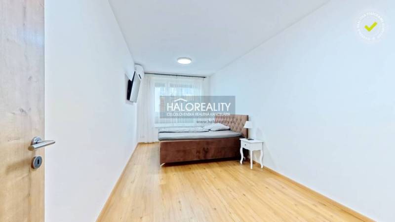 A bright bedroom with a bed, a nightstand, and a TV in a two-room apartment, with a wood-patterned floor.