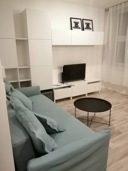 A studio apartment with a gray sofa, television, white furniture, and a wooden-patterned floor.