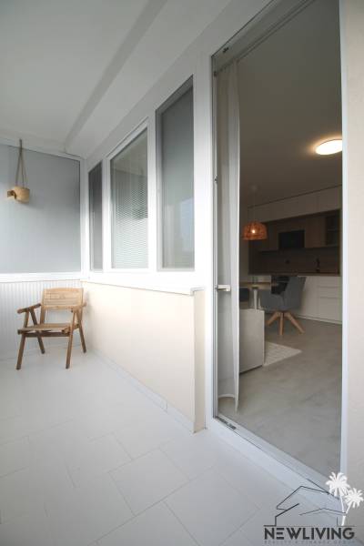 A furnished balcony with a wooden chair leads into a studio apartment through glass doors.