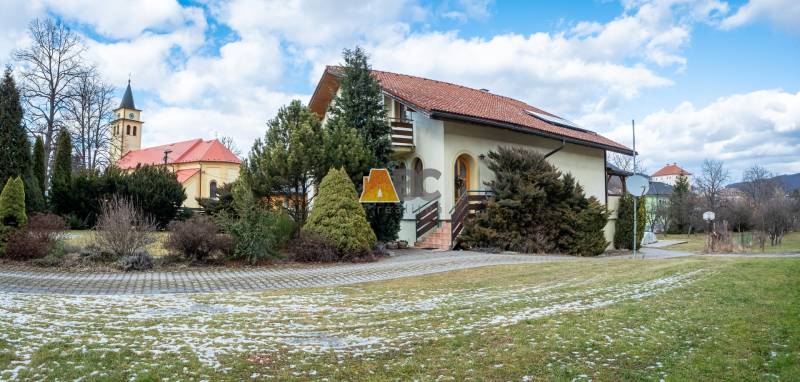 A villa on Andreja Sládkoviča Street in Žarnovica, surrounded by greenery, with a church in the background.