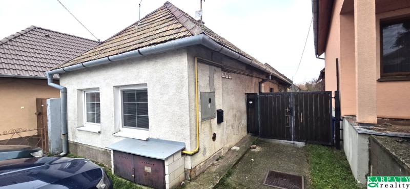 Family house in Dolné Orešany, gray plaster, two windows, sloped roof, adjacent cars.