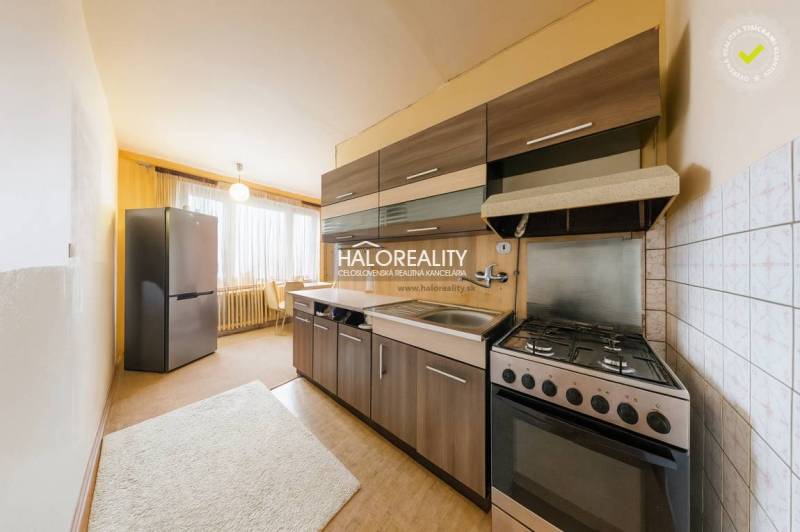 The kitchen of a three-room apartment with dark cabinets, light walls, and a large window.
