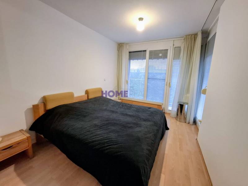 Bedroom in a 2-room apartment with wood-patterned flooring and large windows.
