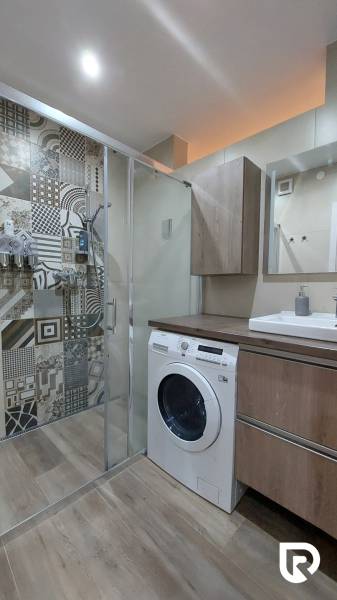 A bathroom with a washing machine, a shower, and a floor with a wooden decor in a 3-room apartment.