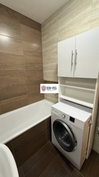 Bathroom in a studio apartment with a bathtub, washing machine, and floor with wooden decor.