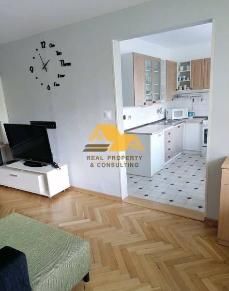 Living room in a three-room apartment with a wooden decor floor and a kitchen in the background.