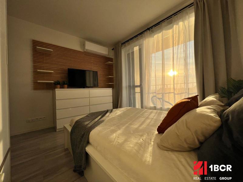 Bedroom in a one-room apartment with wood-patterned flooring and a view of the balcony.