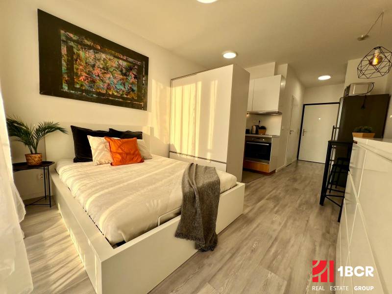 Interior of a studio apartment with a bed, kitchen, and wooden decor flooring.