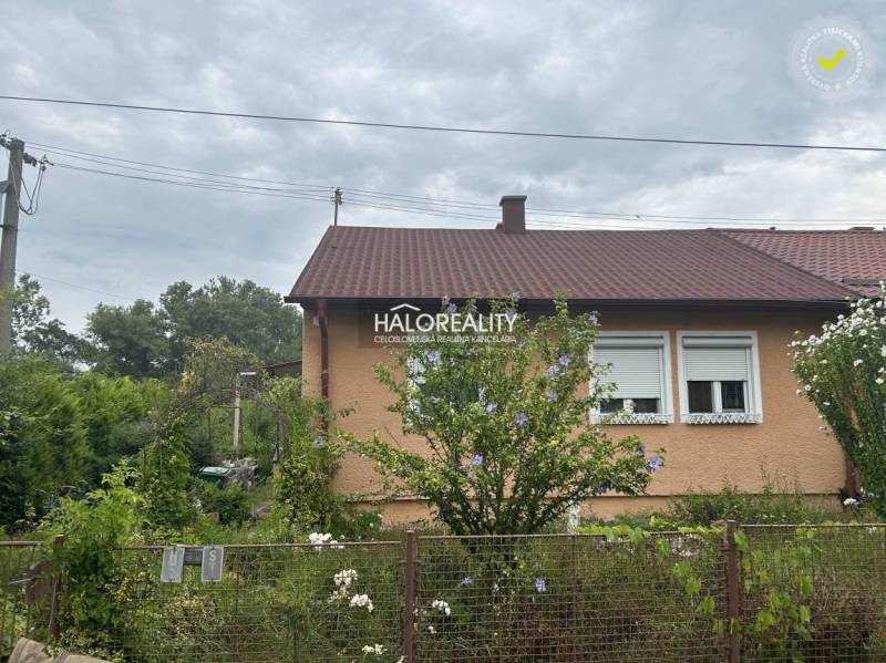 A family house with a red roof surrounded by greenery, flowers, and a fence.