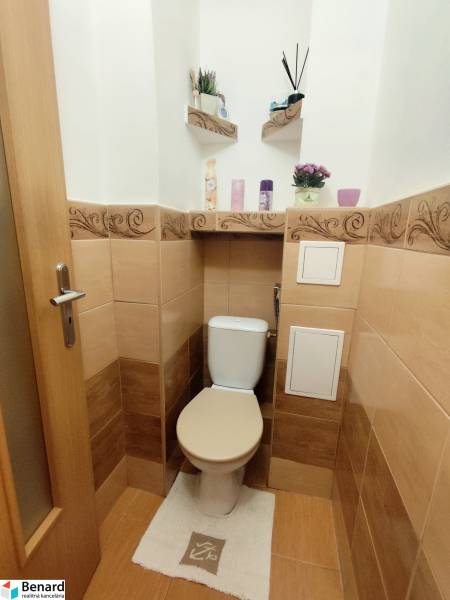A toilet in a 3-room apartment with beige tiles and decorative shelves.