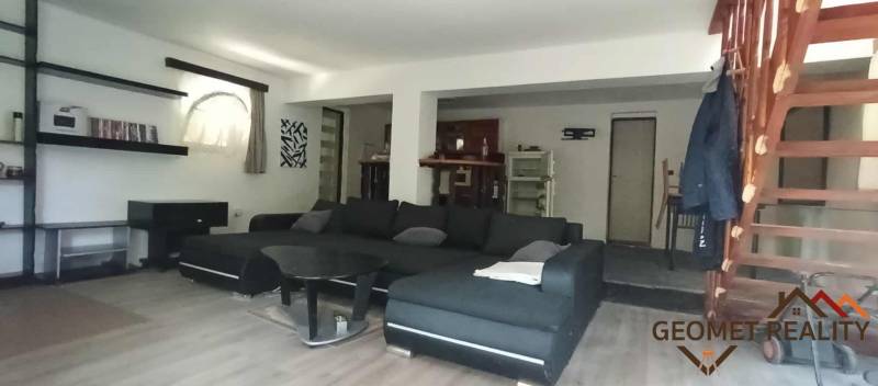 The living room of the cottage with a sofa, a table, and stairs, on a floor with a wooden decor.