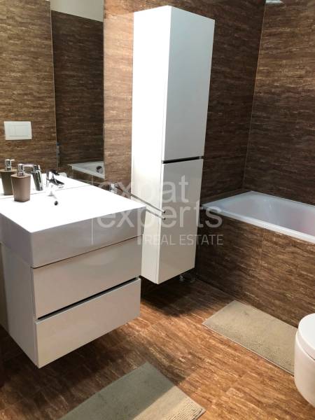 A bathroom in a 2-room apartment with brown tiles, white furniture, and a bathtub.