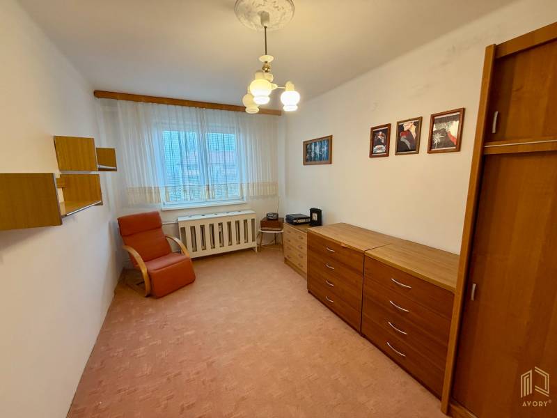 A room in a 3-room apartment with a carpet, a chest of drawers, an armchair, and wall shelves.