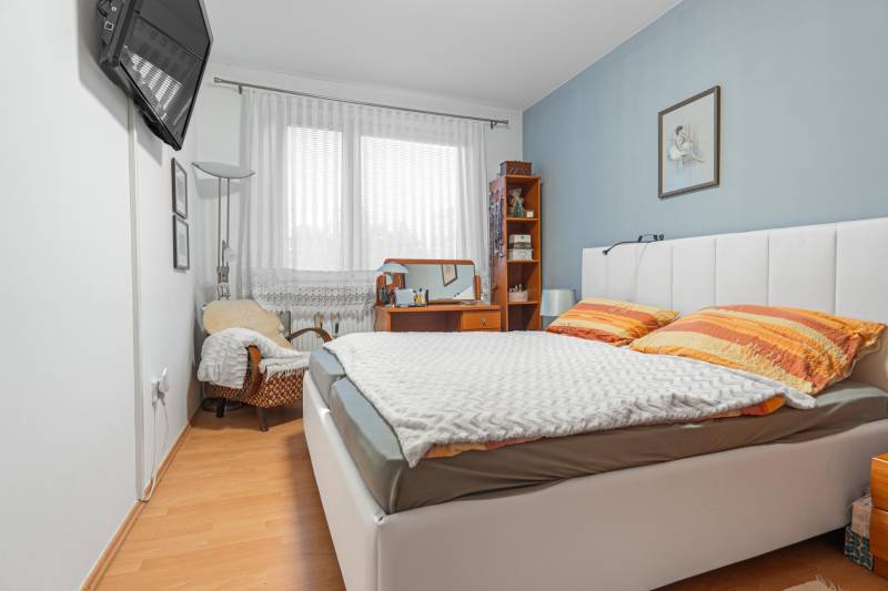 A bedroom with a bed, chair, and table in a 4-room apartment with a wood-patterned floor.