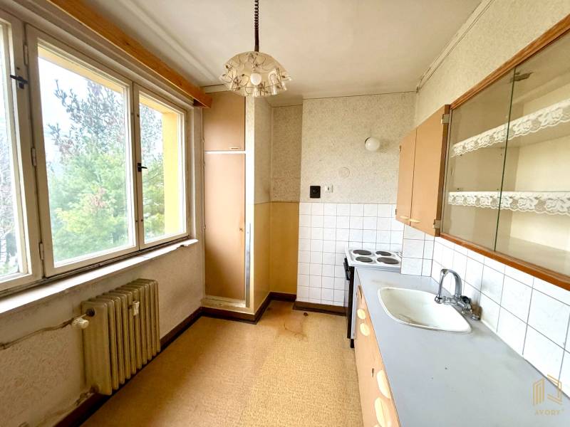 A kitchen in a 2-room apartment with a large window, older furniture, and a light wall.