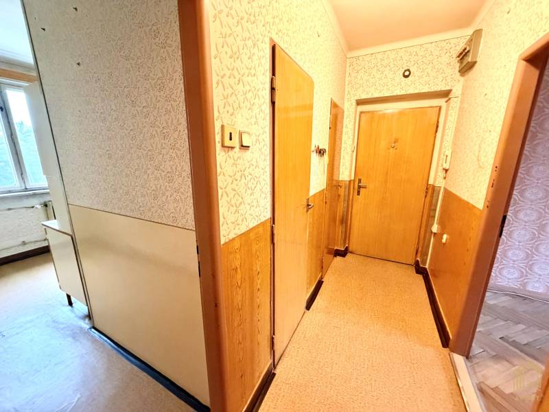 A hallway in a 2-room apartment with light wallpaper, doors, and a window on a floor with a wooden decor.
