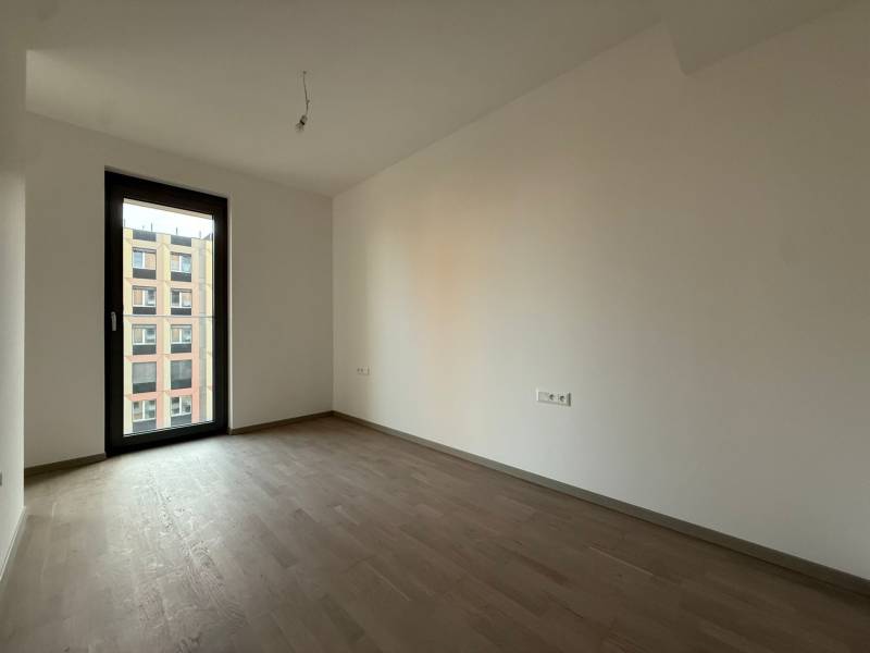 An empty room with wood-patterned flooring in a two-room apartment, large window with a view.